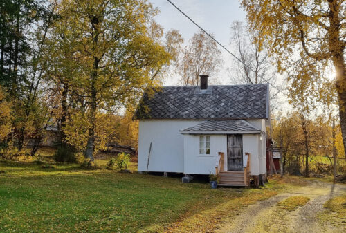 Et lite, hvitt trehus med takspon og en liten veranda, omgitt av høsttrær og en gresskledd hage under en klar himmel.