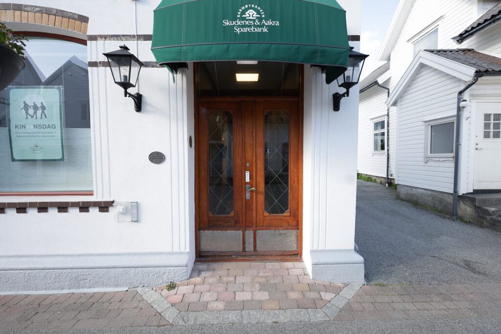 En dobbeltdør i tre med glassfelt, plassert i en hvit bygning under en grønn markise med påskriften Skudenes & Aakra Sparebank. To vegglamper flankerer inngangen.