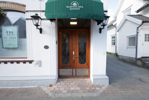 En dobbeltdør i tre med glassfelt, plassert i en hvit bygning under en grønn markise med påskriften Skudenes & Aakra Sparebank. To vegglamper flankerer inngangen.