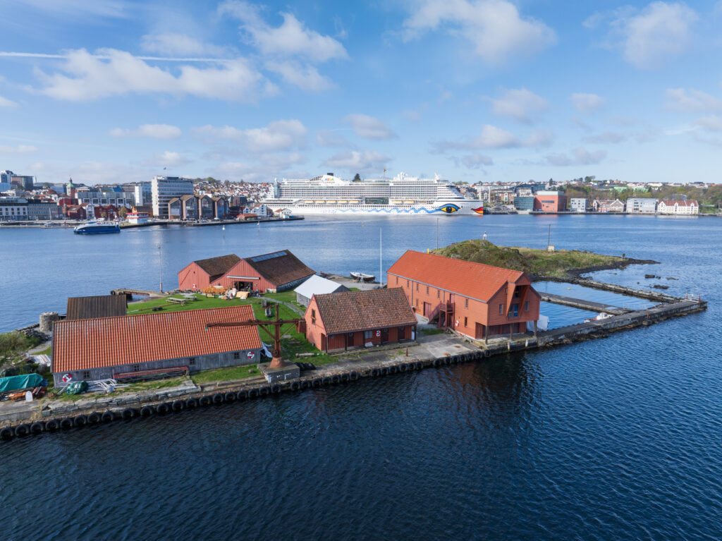 En klynge bygninger med røde tak på en liten øy nær kysten, med et stort cruiseskip og et bylandskap synlig i bakgrunnen under en blå himmel.