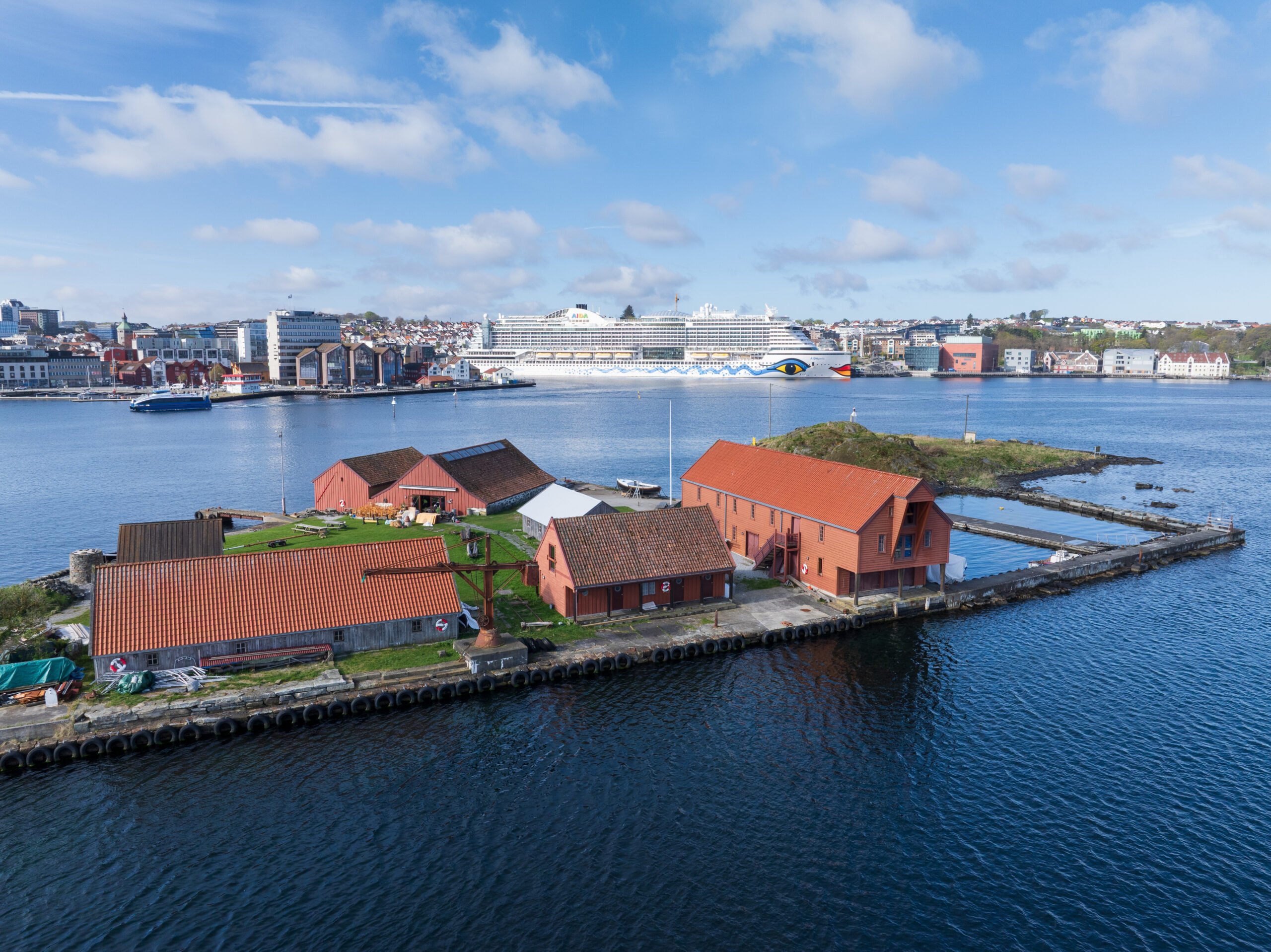 En klynge bygninger med røde tak på en liten øy nær kysten, med et stort cruiseskip og et bylandskap synlig i bakgrunnen under en blå himmel.