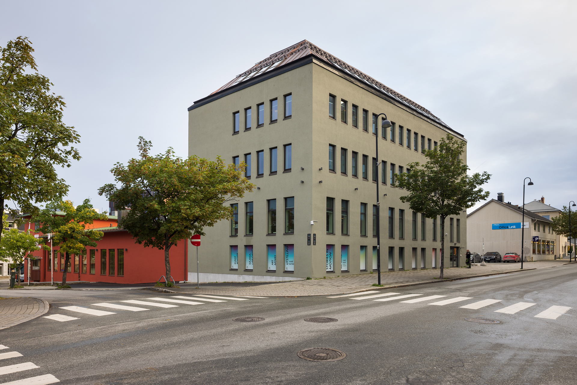 En moderne fireetasjes bygning med mange vinduer står på et hjørne, ved siden av en mindre rød bygning, langs en stille gate med fotgjengerfelt og trær.