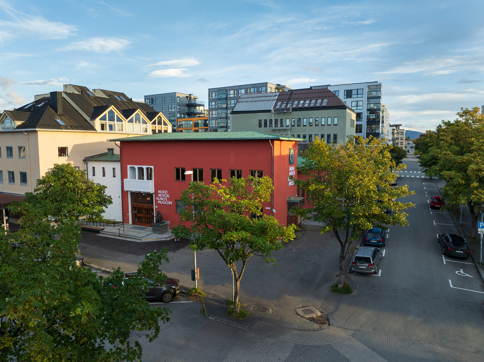 På hjørnet av en stille bygate ligger en rød bygning med navnet Nord Norsk Kunstmuseum, omgitt av trær og moderne bygårder under en delvis overskyet himmel.