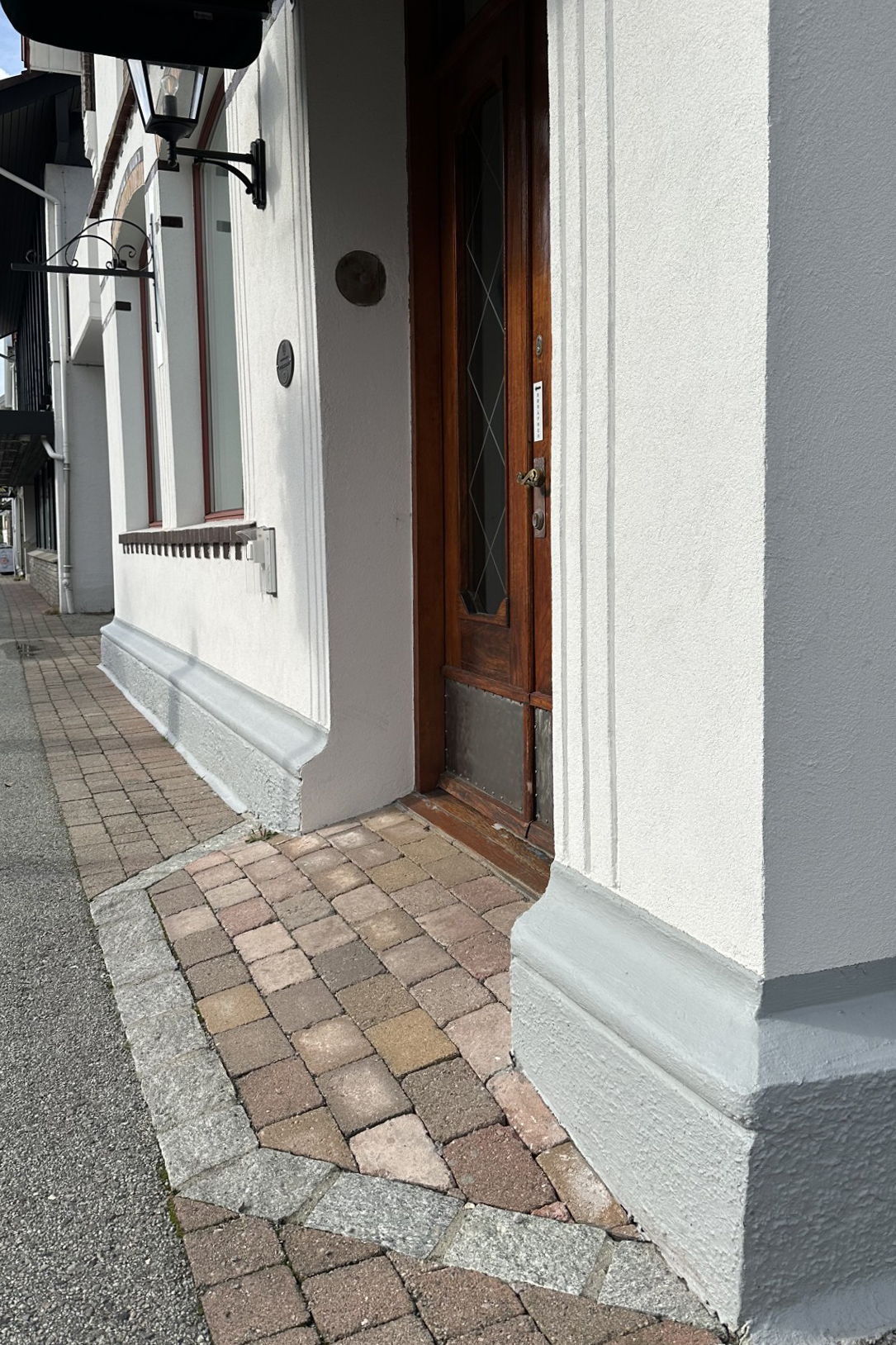 En tredør i en hvit bygning med et vindu ved siden av, omkranset av lysegrå listverk og en gangvei av belegningsstein.