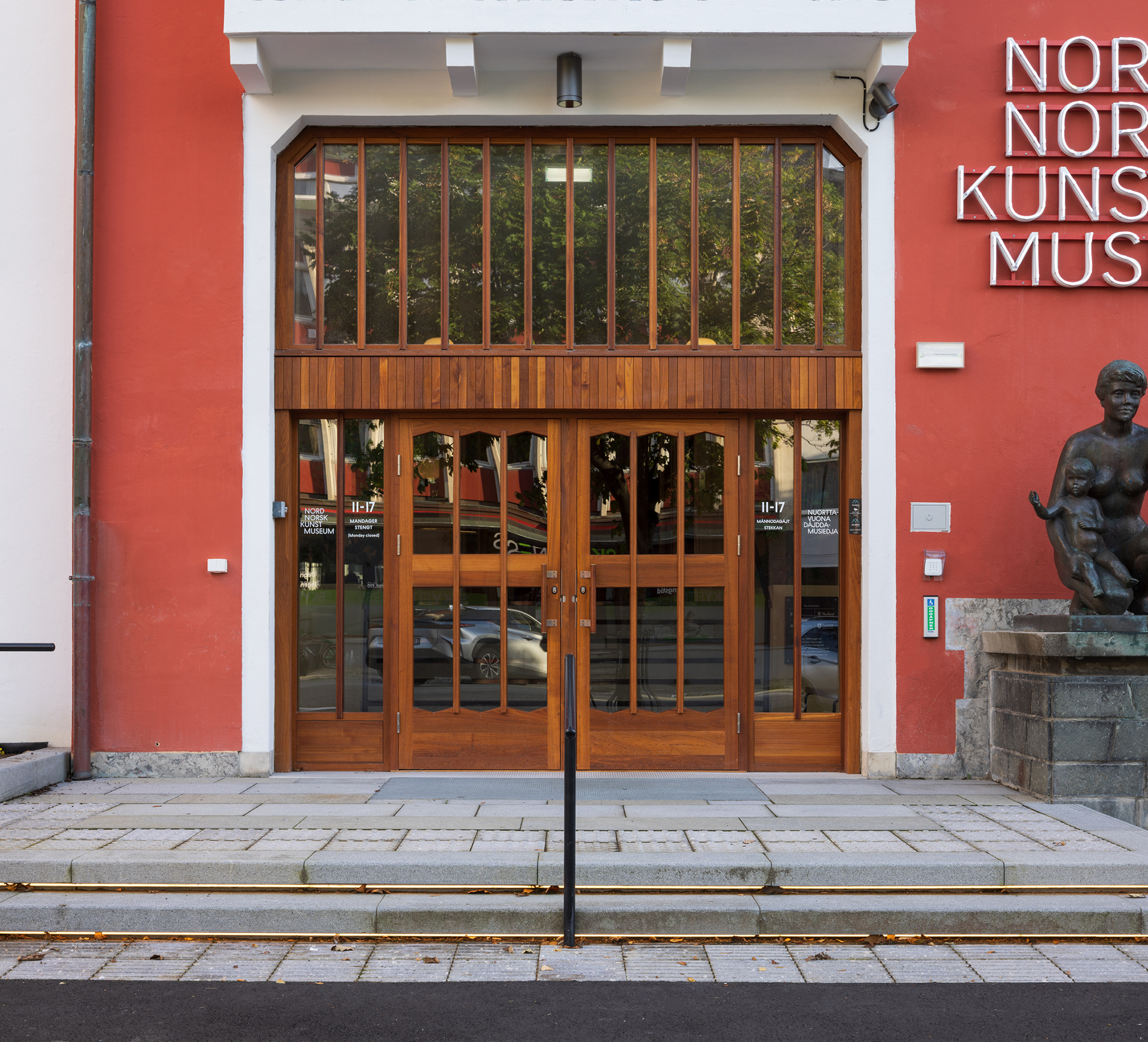 Inngangsdører i glass og tre til en rød bygning med et hvitt skilt med teksten NORSK KUNST MUSEUM og en bronsestatue til høyre.