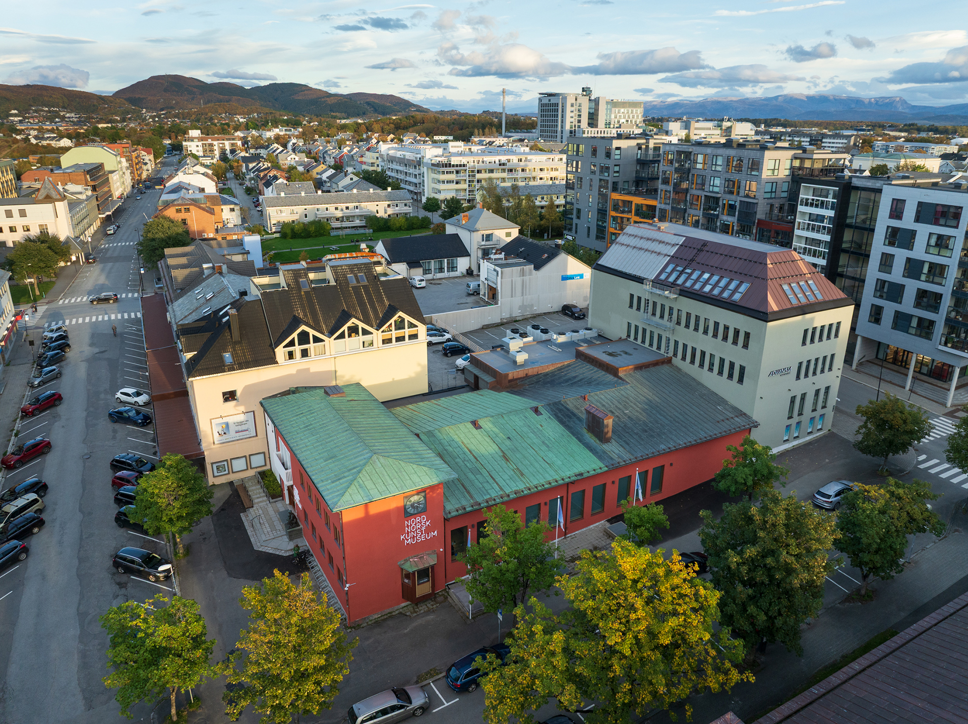 Flyfoto av et moderne byområde med bolig- og næringsbygg, parkerte biler langs gatene og grønne trær i forgrunnen.