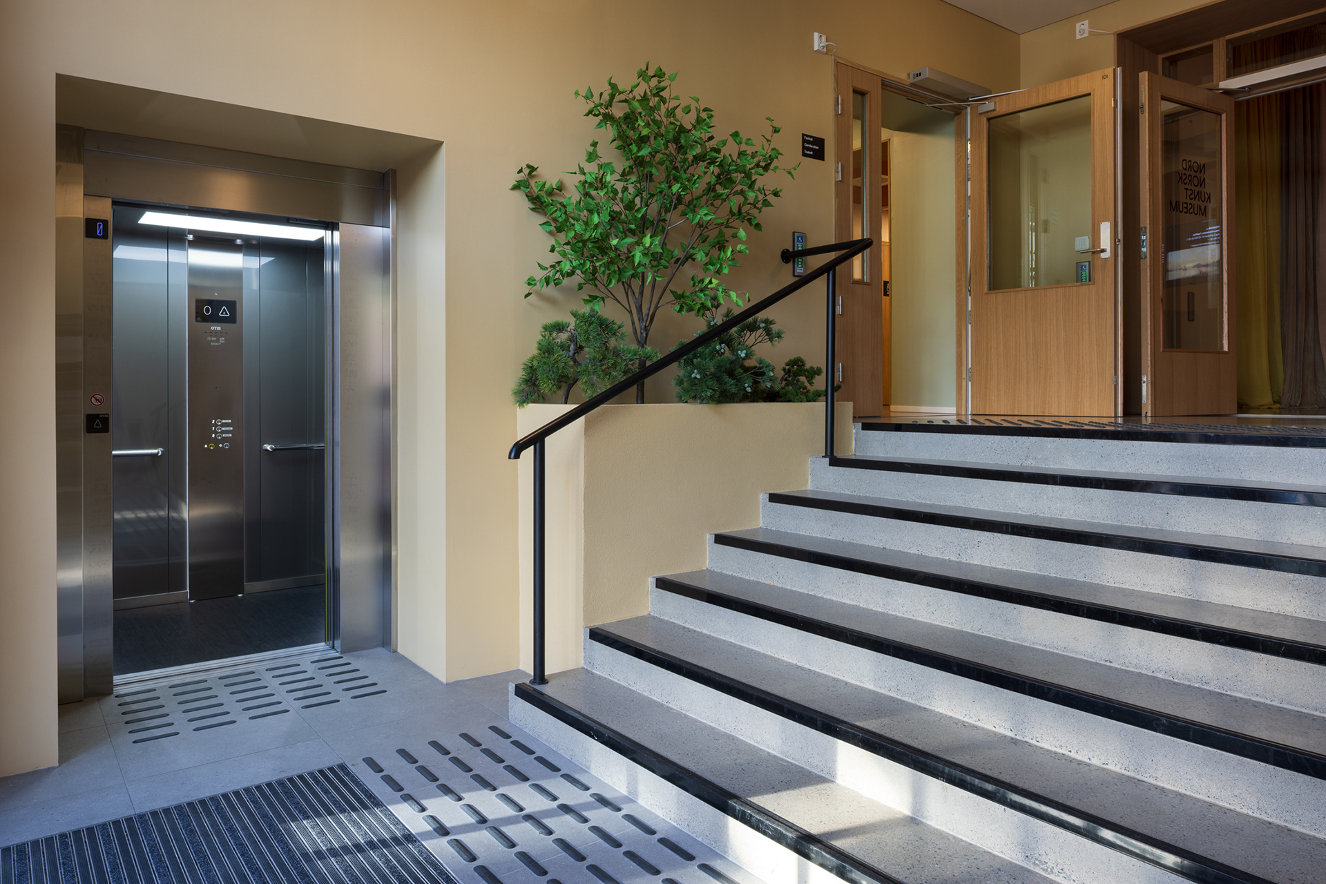 Et moderne innendørsrom med en åpen heis til venstre, trapper med svart rekkverk til høyre og et tre i potte i midten, nær doble tredører.