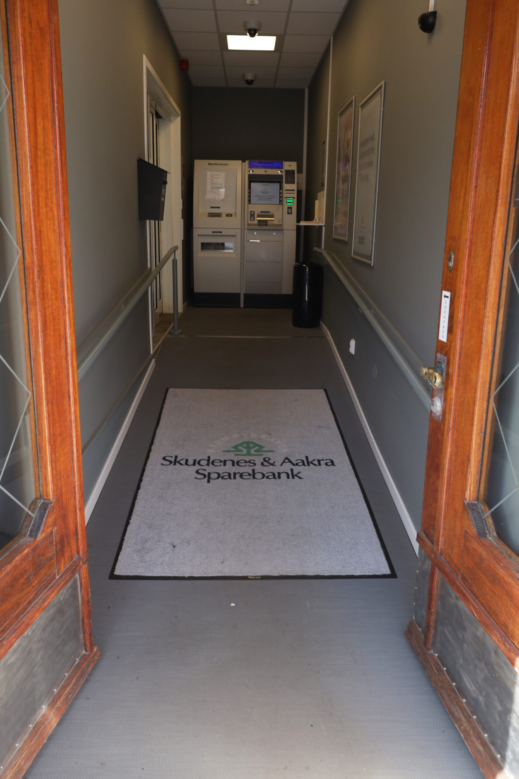 Inngangsparti som leder til minibankene, med en gulvmatte med Skudenes & Aakra Sparebanks logo. Veggene er grå, og plakater er synlige på høyre side.