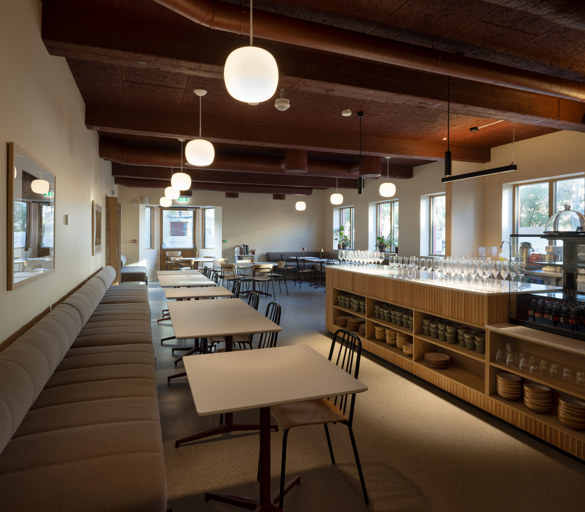 En romslig, moderne spiseplass med sirlig oppstilte bord og stoler, pendellamper, store vinduer og en buffetdisk med glass og tallerkener.