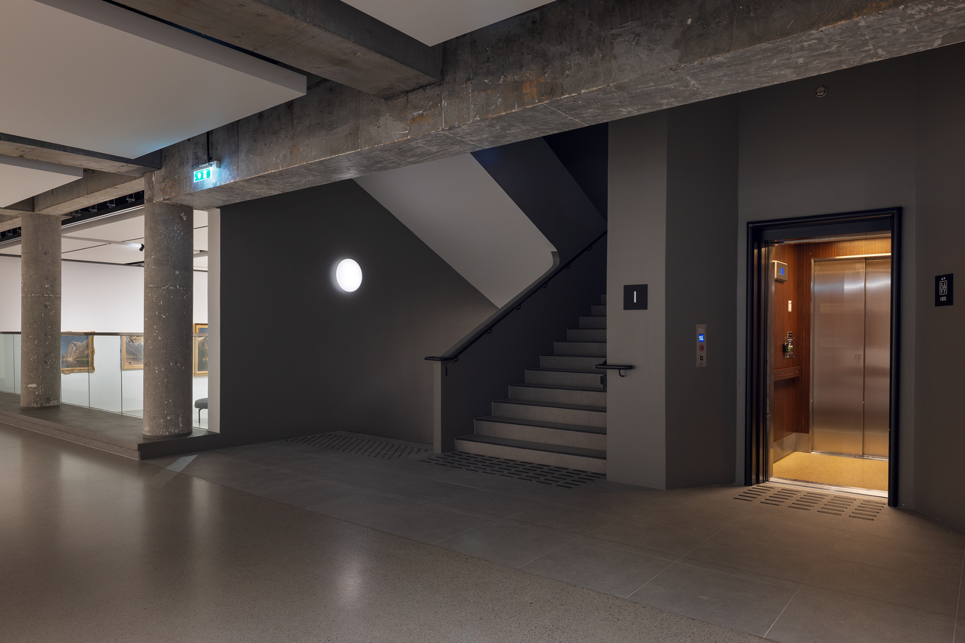 Et moderne bygningsinteriør med betongbjelker, en trapp og en åpen heis ved siden av en korridor.
