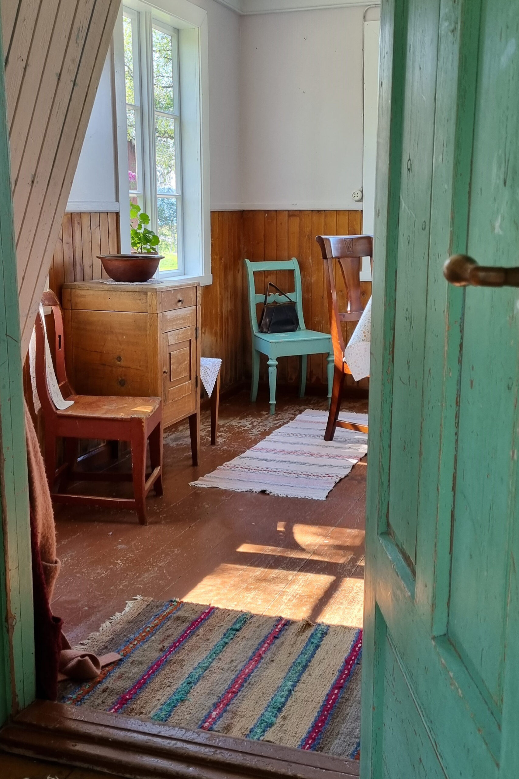 Et blikk gjennom en turkis dør inn i et solfylt rom med tremøbler, en potteplante på et skap, stripete tepper og et vindu som slipper inn naturlig lys.