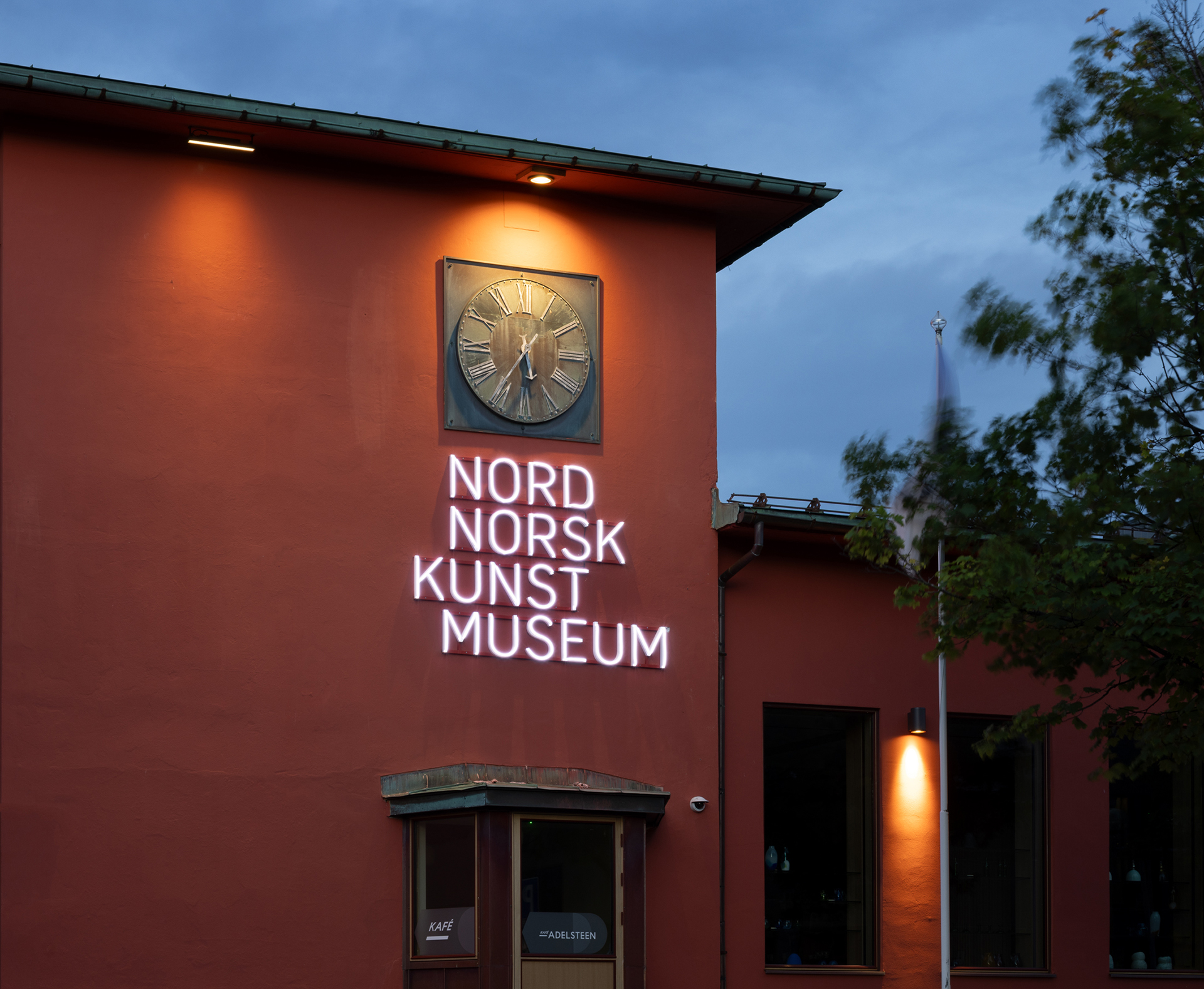 En rød bygning med en opplyst klokke og et neonskilt med teksten Nord Norsk Kunstmuseum over en døråpning, fotografert i skumringen.