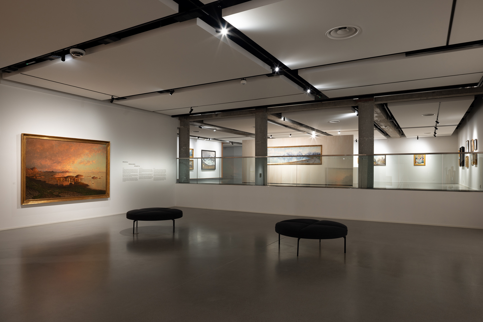 Moderne kunstgalleriinteriør med innrammede landskapsmalerier på hvite vegger, svarte benker, polert betonggulv og spotlights i taket.