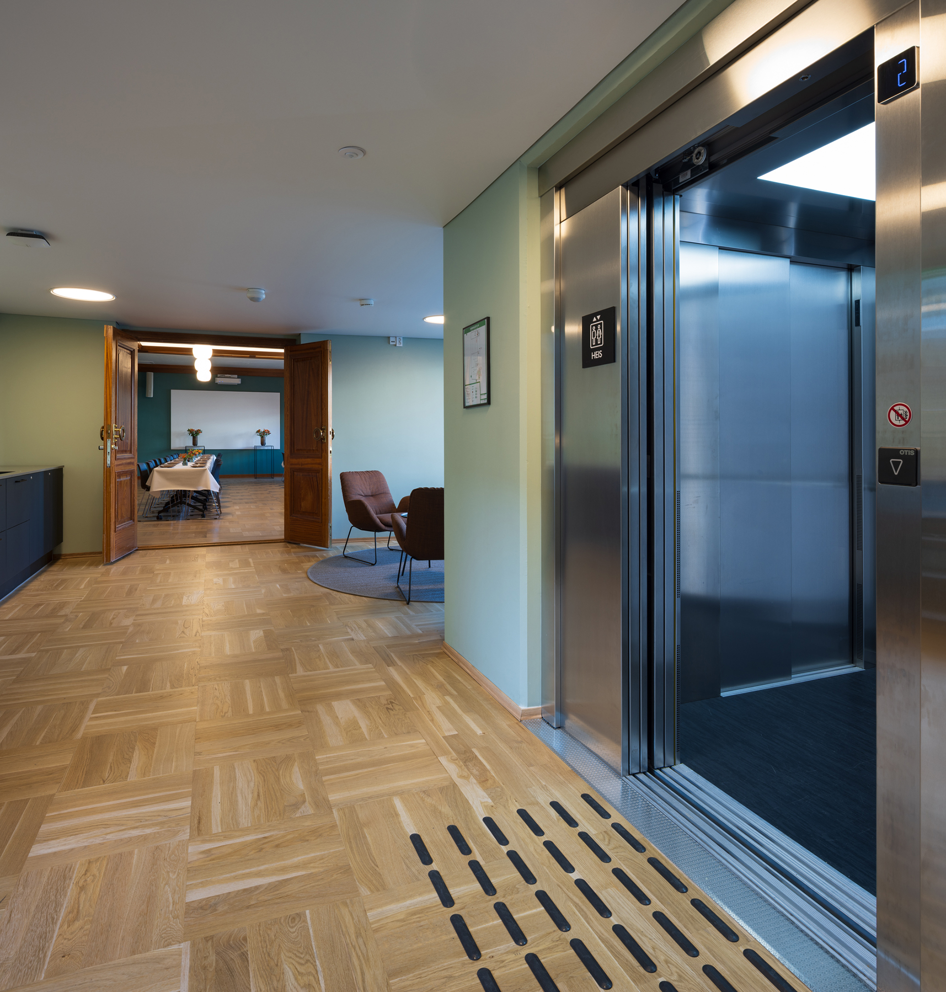 En moderne bygning med en åpen heis på høyre side, parkettgulv og en gang som fører til et rom med bord og stoler.