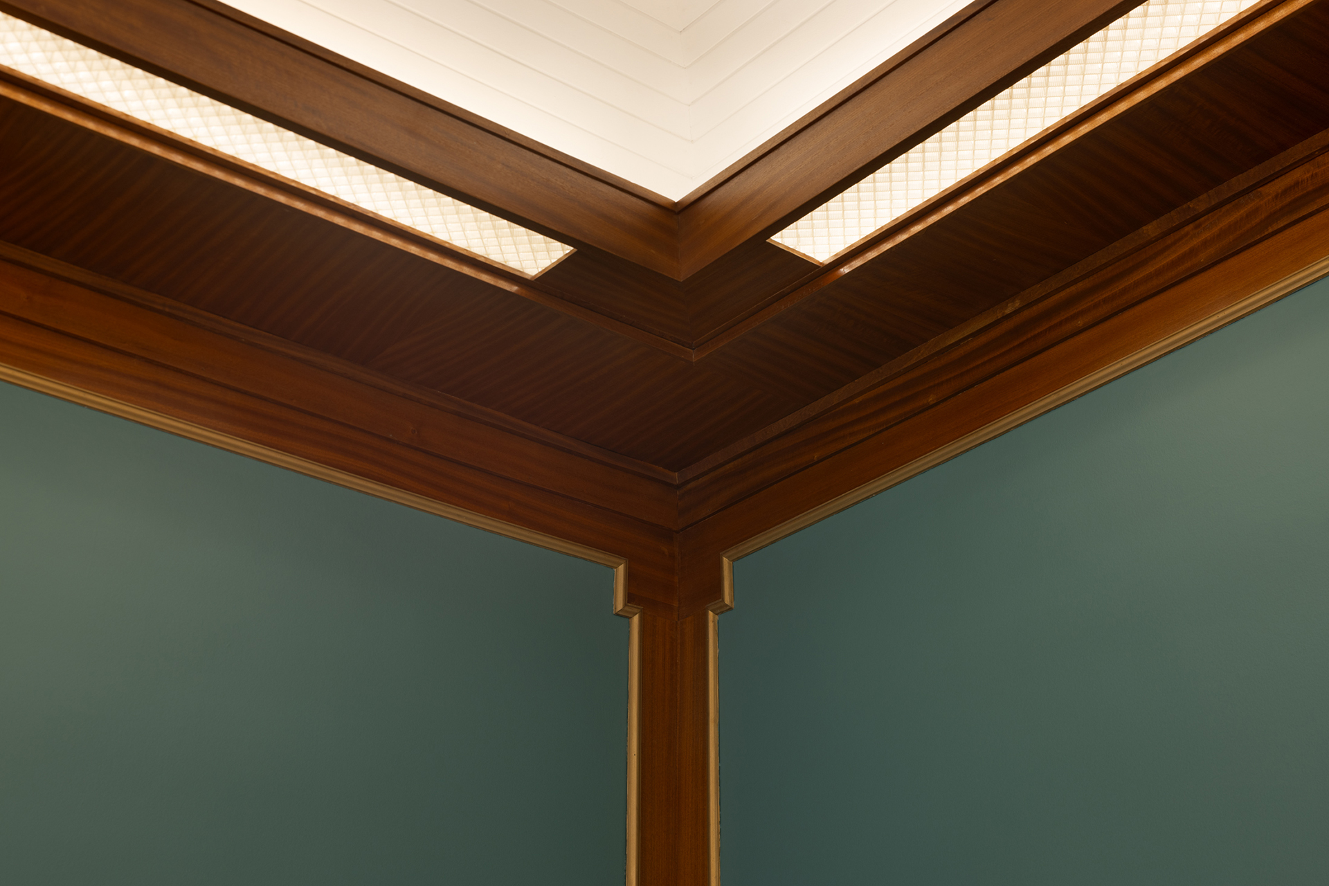 Nærbilde av et romhjørne med grønne vegger, trelister og et tak med innfelt belysning og geometriske mønstre.