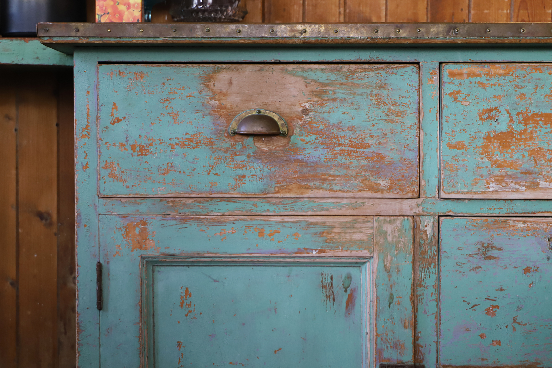 Nærbilde av et gammelt, forvitret, turkist treskap med avskallet maling og messingbeslag mot en trevegg.