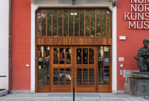 Et moderne inngangsparti i tre og glass til en bygning med røde vegger, en statue til høyre og et museumsskilt delvis synlig over.