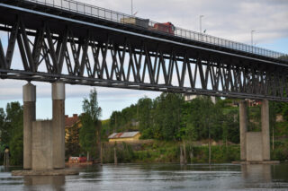 En fagverksbro i metall med en lastebil på toppen spenner over en elv, støttet av betongpilarer, med bygninger og trær i bakgrunnen.