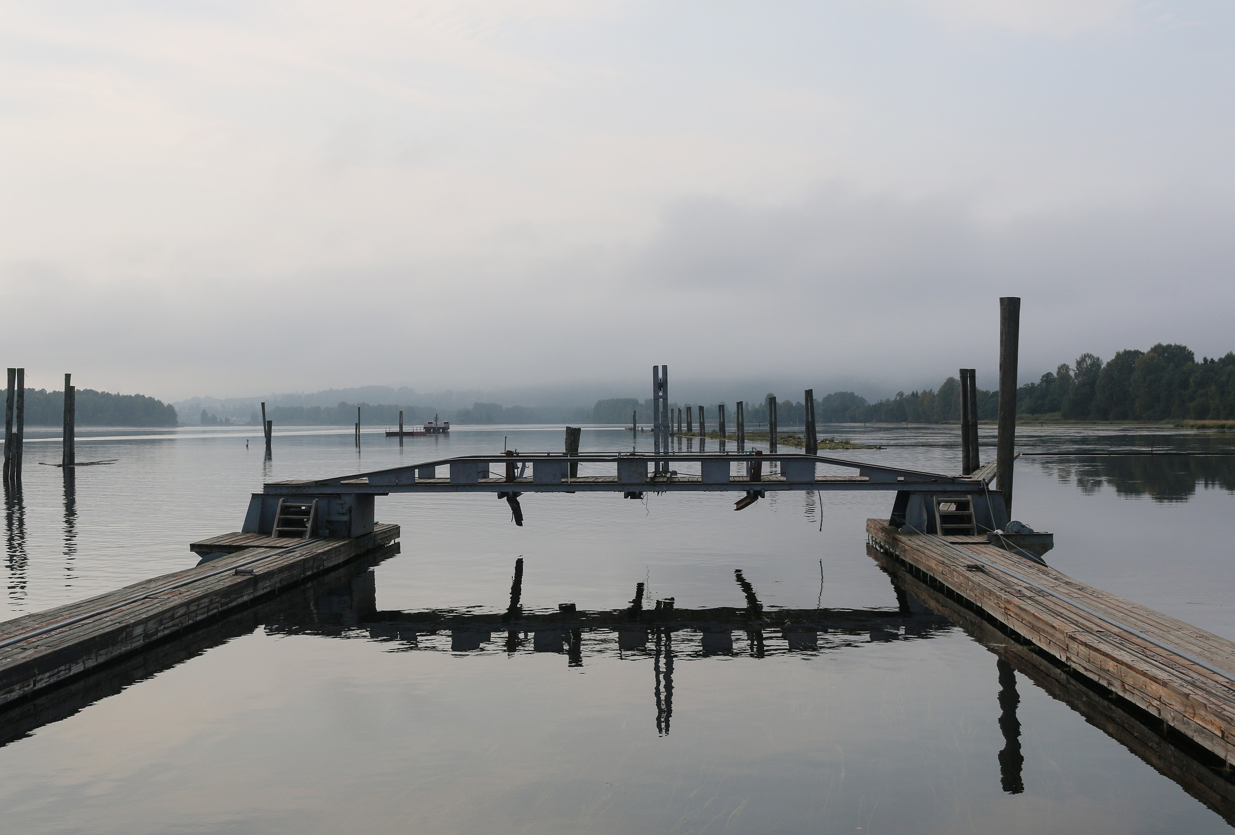 En trebrygge strekker seg ut i en rolig innsjø med stolper og en liten brokonstruksjon, omgitt av trær og en skyet himmel i bakgrunnen.
