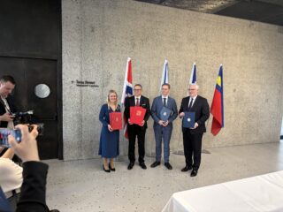 Fire personer står foran nasjonale flagg med offisielle dokumenter i hendene, mens fotografer tar bilder i en formell innendørs setting.