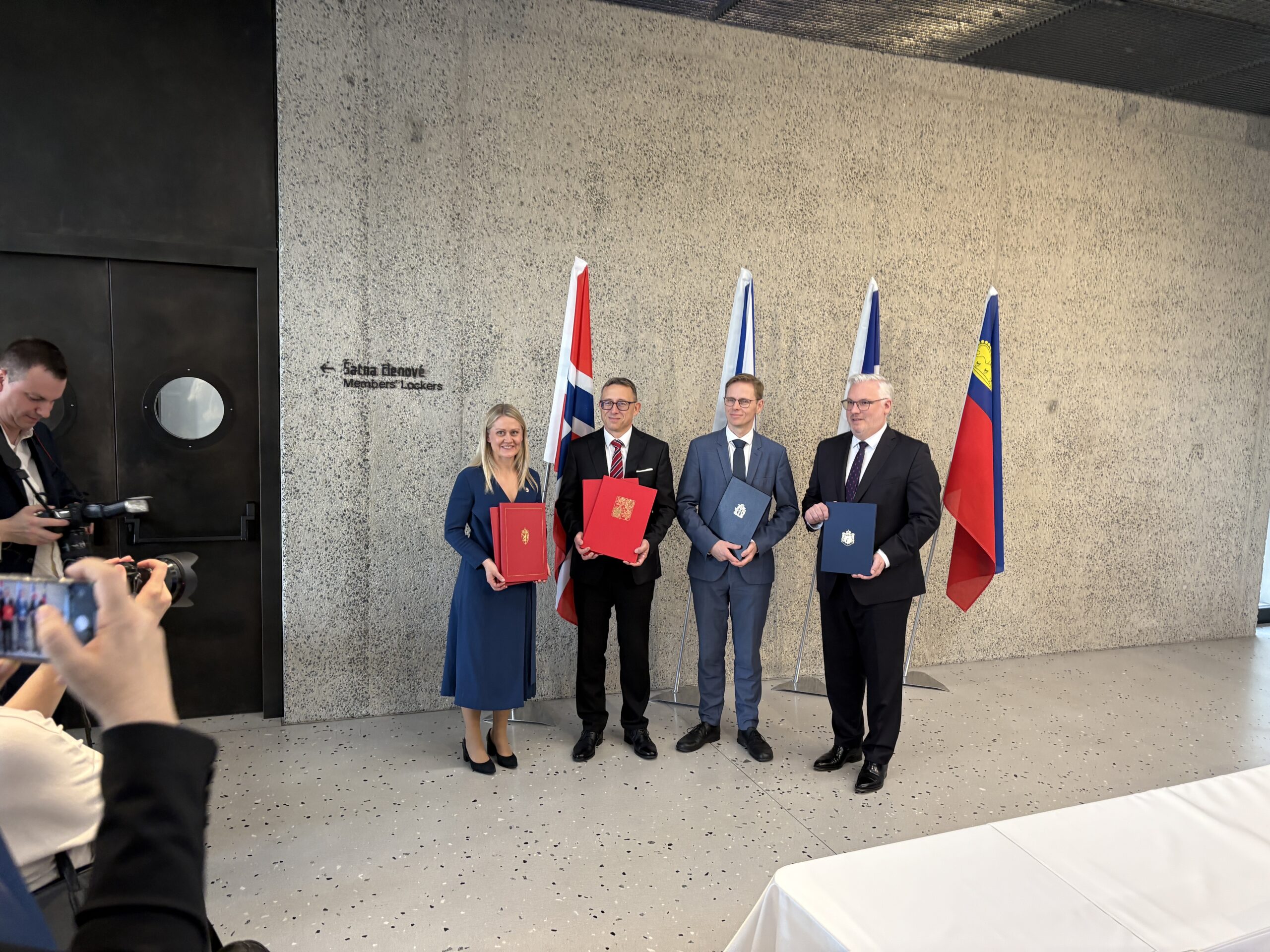 Fire personer står foran nasjonale flagg med offisielle dokumenter i hendene, mens fotografer tar bilder i en formell innendørs setting.