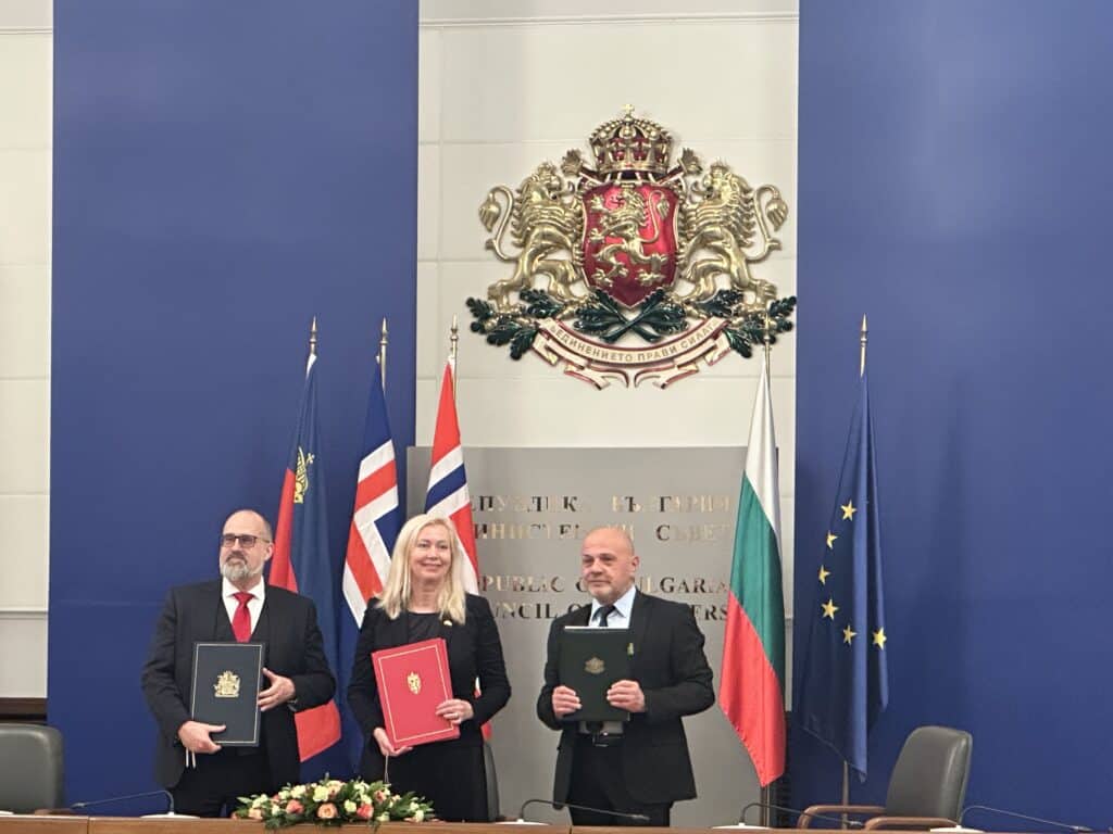Tre personer står med dokumenter i hendene foran et podium, med flere flagg og et nasjonalt emblem på den blå veggen bak dem.