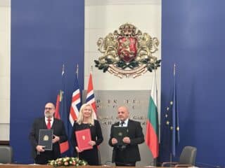 Tre personer står med dokumenter i hendene foran et podium, med flere flagg og et nasjonalt emblem på den blå veggen bak dem.