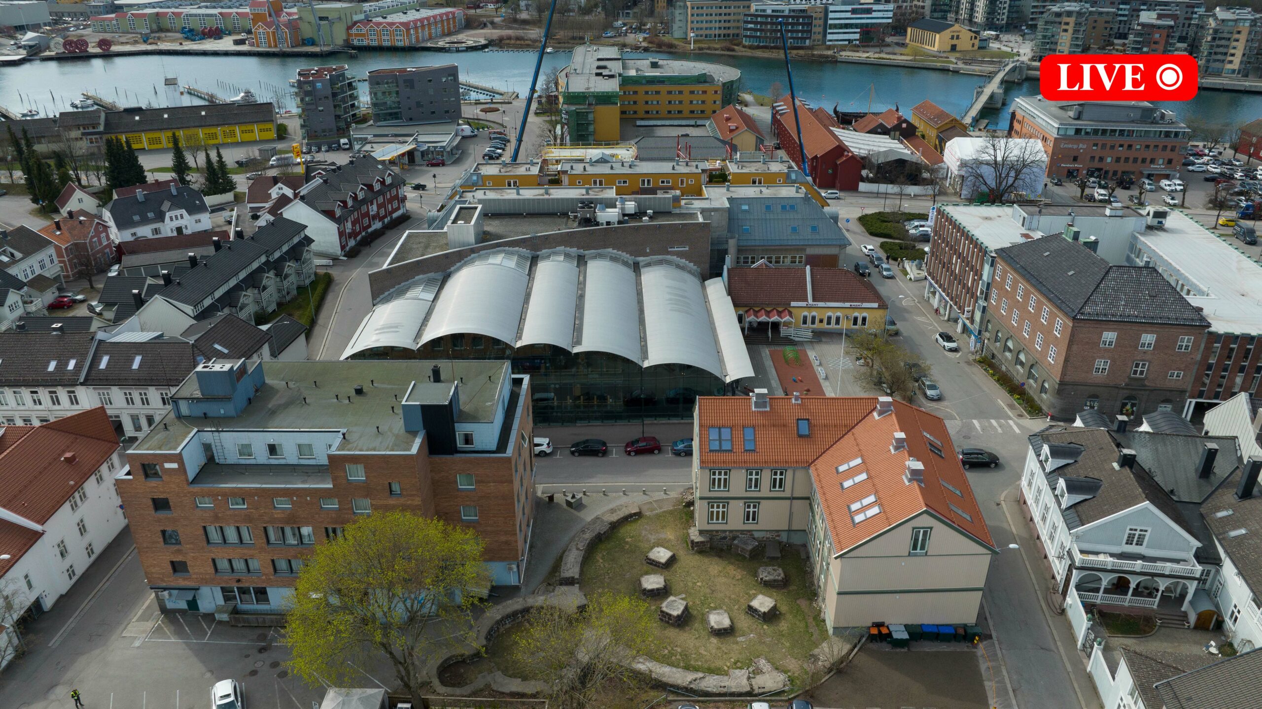 Flyfoto av et lite byområde med moderne og tradisjonelle bygninger, inkludert en vern-inspirert bygning med buet tak i midten, og en elv i bakgrunnen. Direkte-TV-grafikk vises øverst i høyre hjørne.