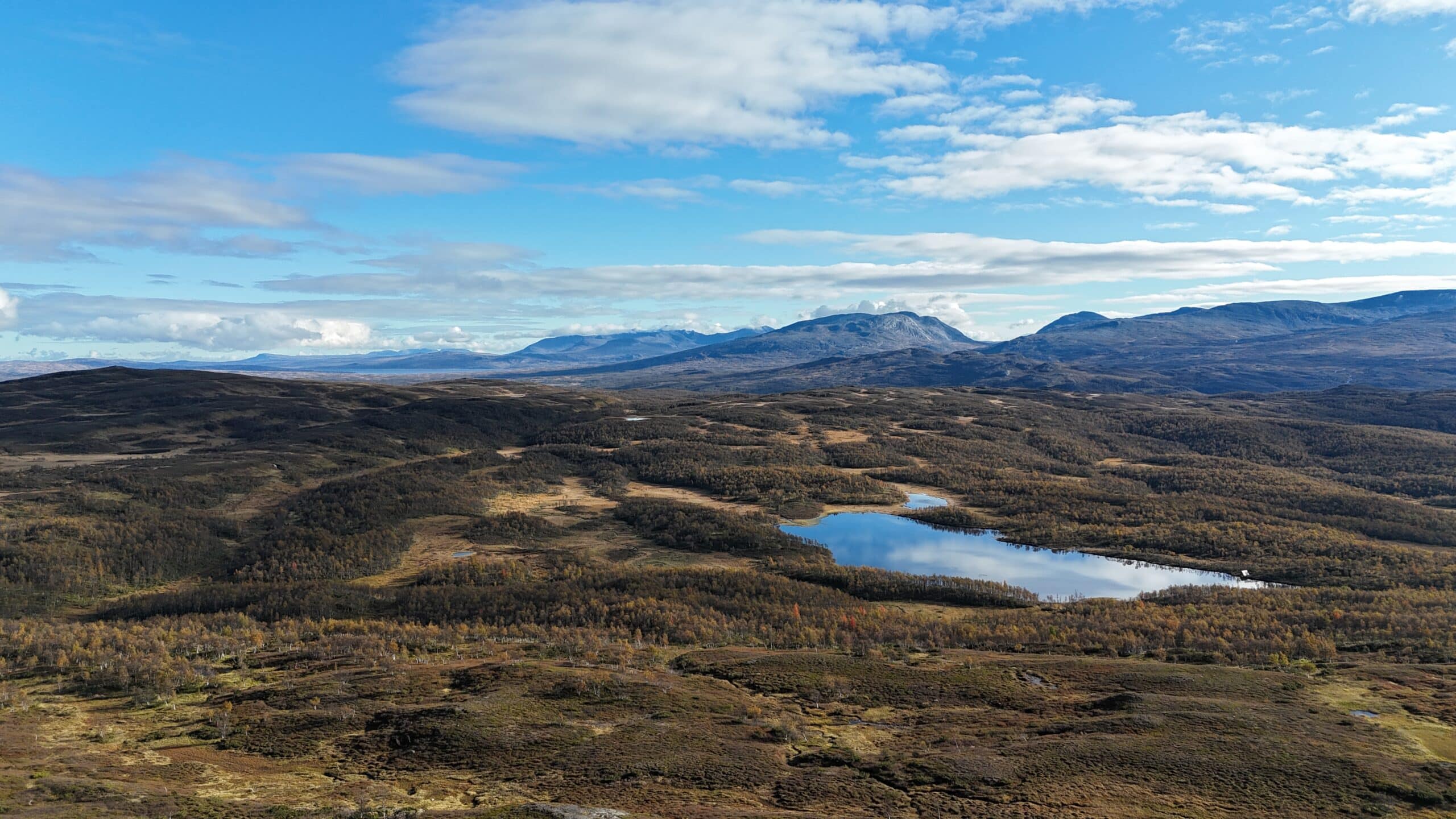 Flyfoto av et kupert landskap med spredte trær, en speilende innsjø og fjell i det fjerne under en delvis overskyet blå himmel.
