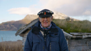 En eldre person iført mørk jakke, hatt med nåler og briller står utendørs foran en rustikk trebygning med fjell i bakgrunnen.