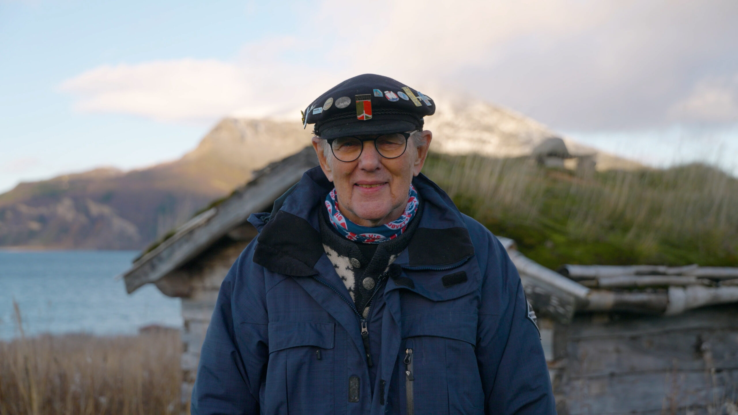 En eldre person iført mørk jakke, hatt med nåler og briller står utendørs foran en rustikk trebygning med fjell i bakgrunnen.