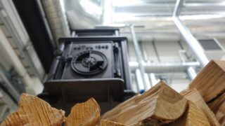 I forgrunnen ligger vedstabler, og i bakgrunnen skimtes en svart vedovn og ventilasjonskanaler av metall.