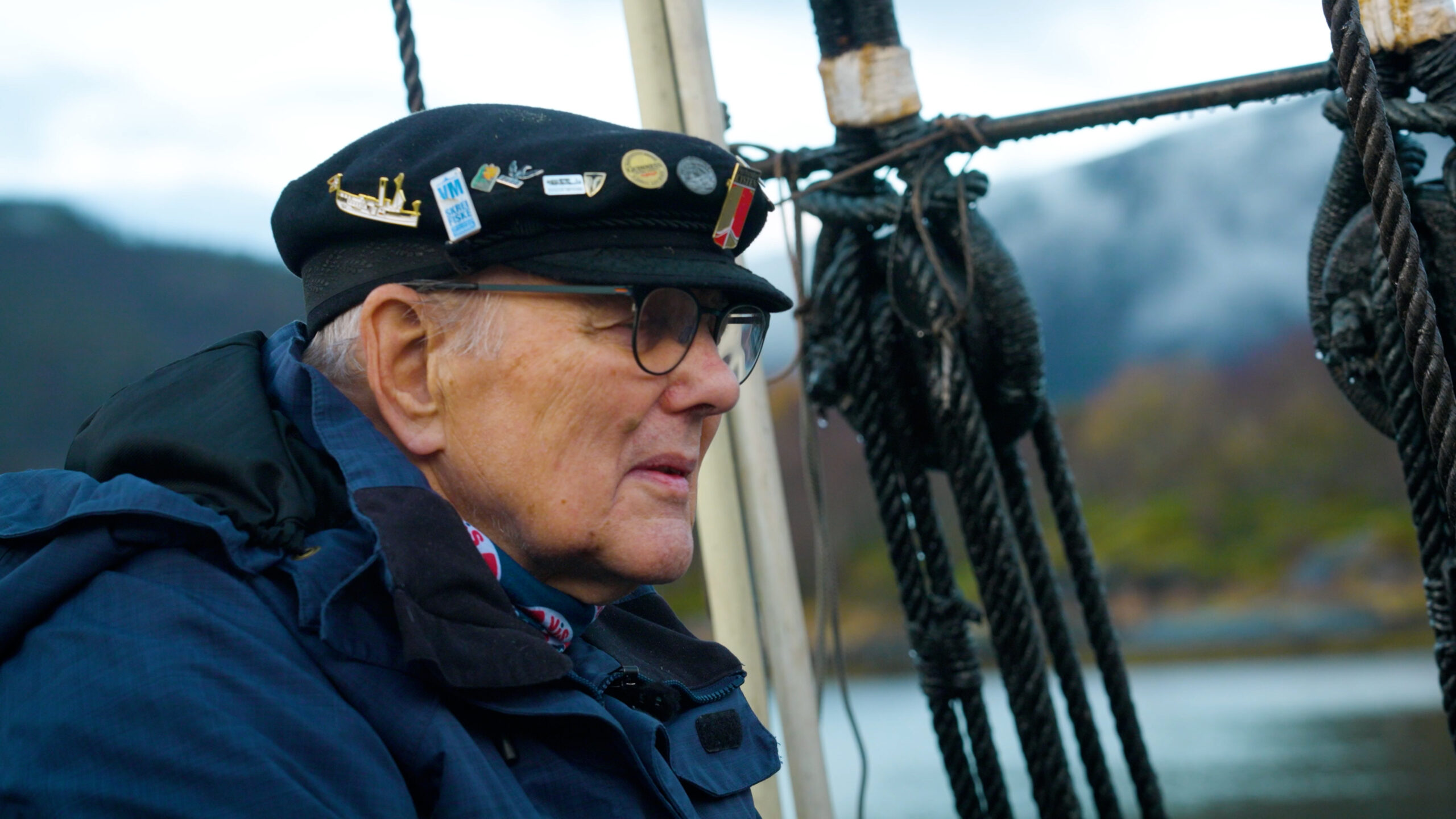 En eldre mann med marinelue med nåler og briller sitter på en båt og ser fremover, med tauverk og et uskarpt landskap i bakgrunnen.