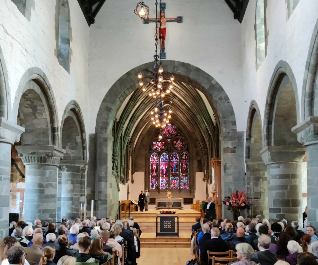 En forsamling sitter inne i Stavanger domkirke, med hvelvede tak, glassmalerier bak alteret og et stort krusifiks som henger fra taket.