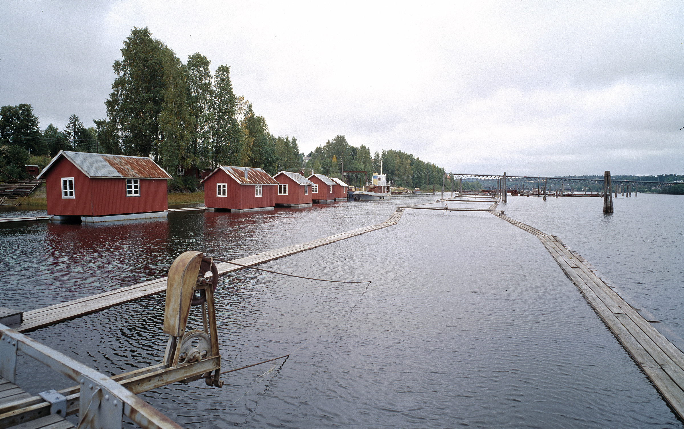 En rekke røde naust av tre langs kanten av en rolig innsjø, med trær i bakgrunnen og en trebrygge som strekker seg ut i vannet under en overskyet himmel.