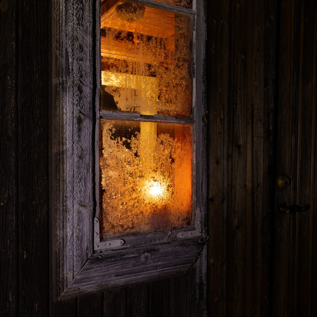 Lillebøle gård i Rollag kommune, i vintermørke, med et frostet vindu hvor det lyser varmt, gult lys fra inne i huset. Lyset er koselig og står i kontrast til mørket utenfor.