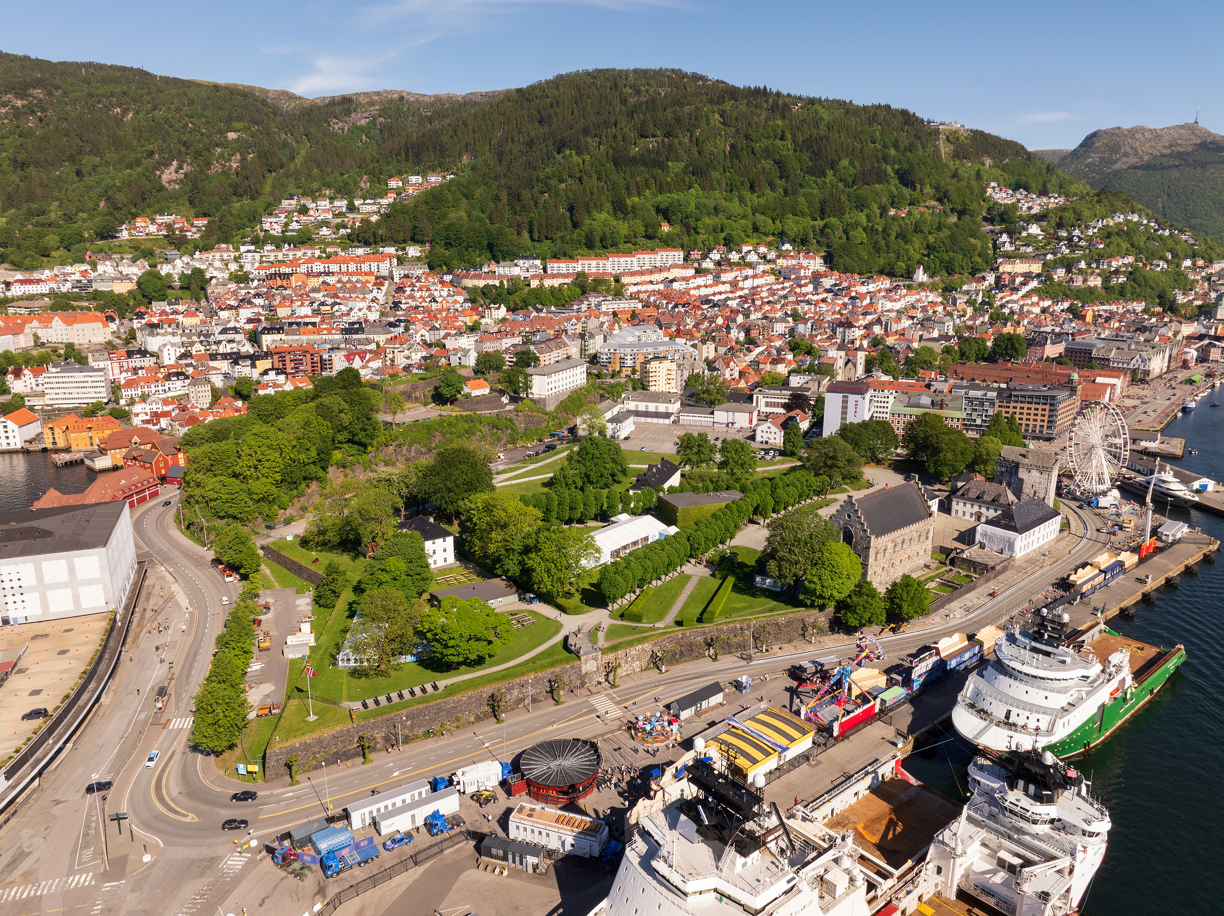 Flyfoto av en kystby med grønne åser, boligblokker, en park, havn, skip og et pariserhjul under en klar himmel.