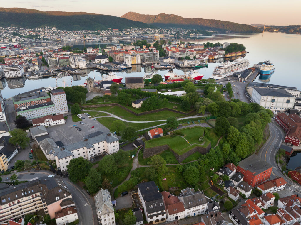 Flyfoto av Bergen, Norge, som viser den gamle festningen, grønne parkområder, havnen med cruiseskip og omkringliggende boligbebyggelse med fjell i bakgrunnen.