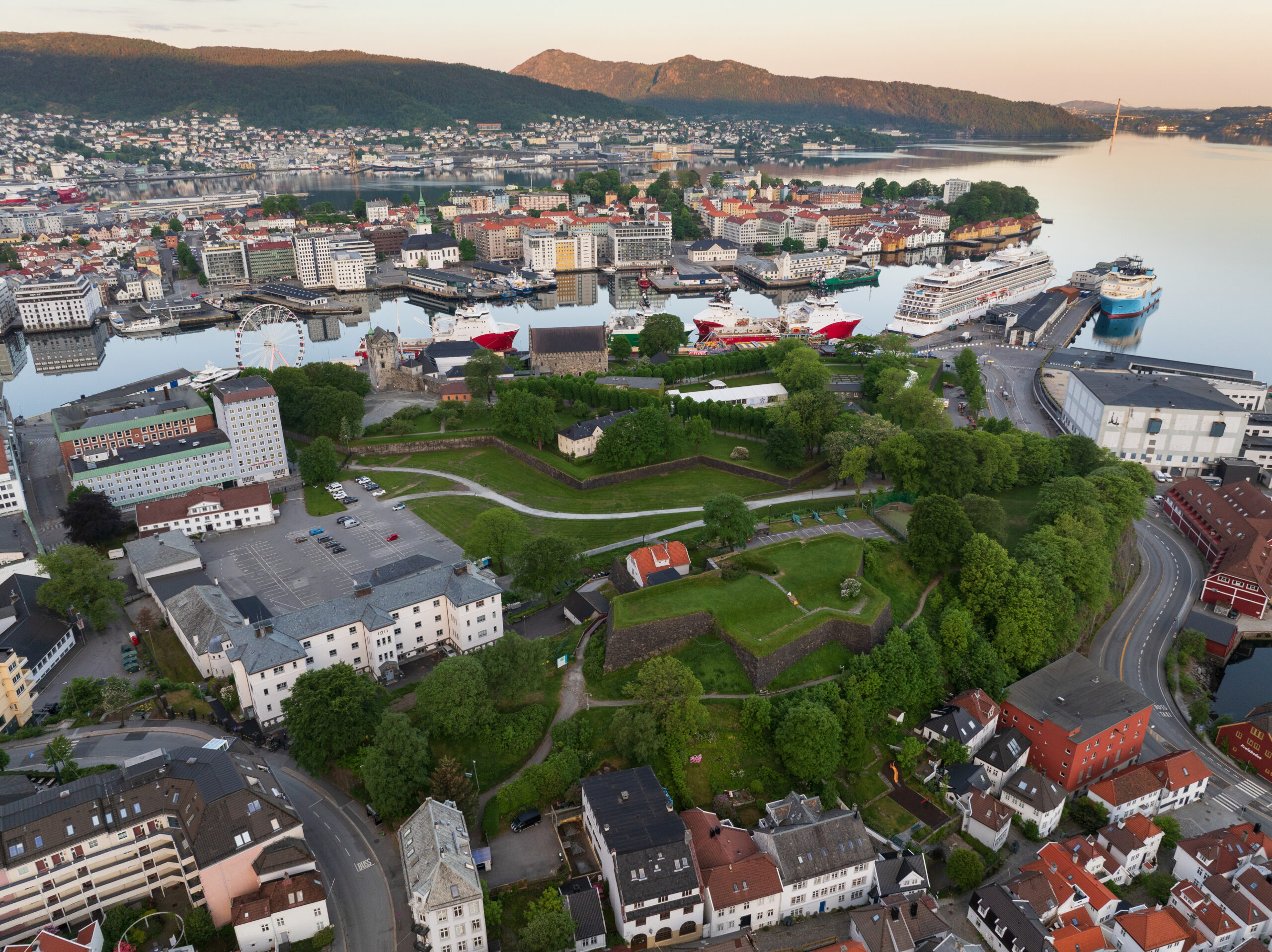 Flyfoto av Bergen, Norge, som viser den gamle festningen, grønne parkområder, havnen med cruiseskip og omkringliggende boligbebyggelse med fjell i bakgrunnen.