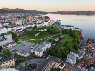 Flyfoto av en kystby med bolig- og næringsbygg, grønne parker og cruiseskip som ligger til kai på rolig vann.