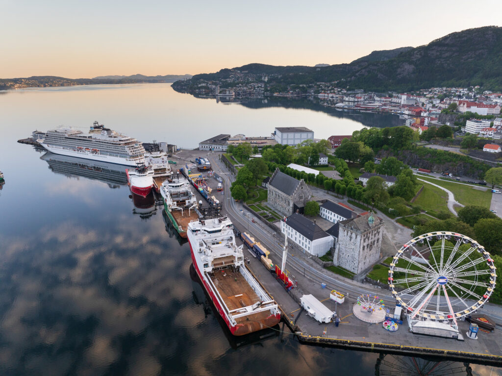 Flyfoto av en havn med skip ved kai, historiske bygninger, et pariserhjul og en kystby omgitt av åser og vann ved soloppgang.