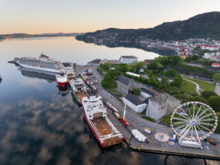Flyfoto av en havn med skip ved kai, historiske bygninger, et pariserhjul og en kystby omgitt av åser og vann ved soloppgang.
