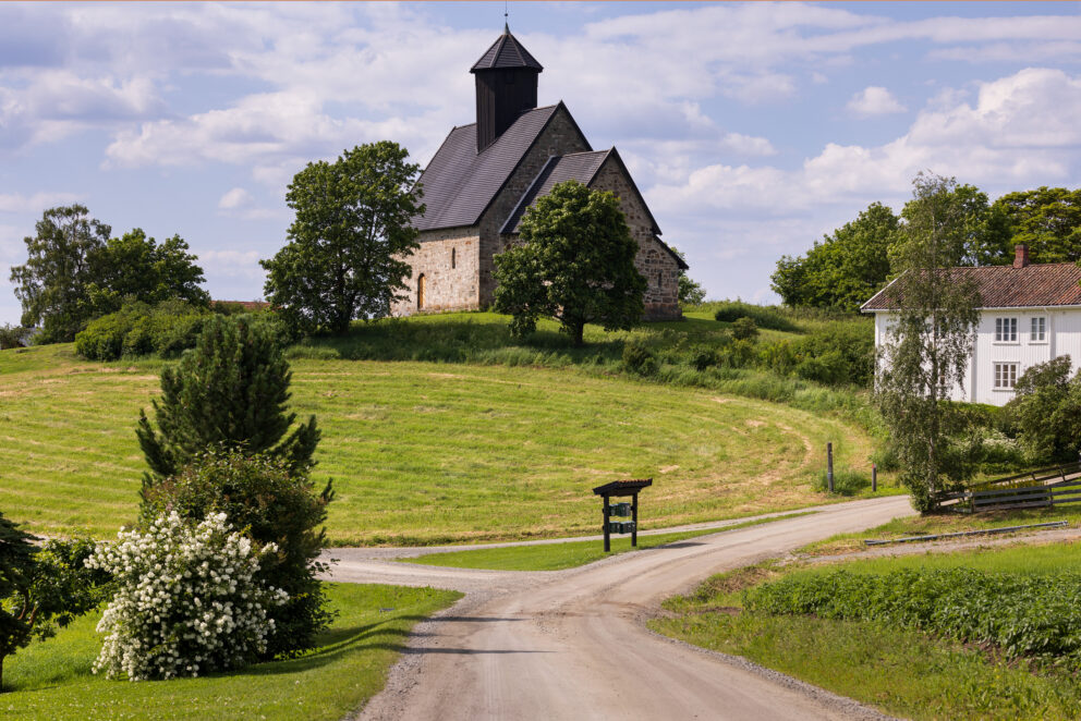 Et landlig landskap med en steinkirke på en gressbakke, trær rundt kirken, et hvitt hus i nærheten og en grusvei som deler seg ved et veiskille i forgrunnen.