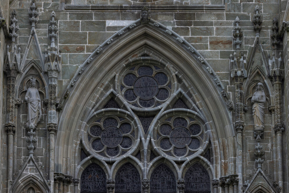 Detaljbilde av en gotisk katedralfasade med spissbuer, utsmykkede sprosser, glassmalerier og steinstatuer plassert i nisjer.