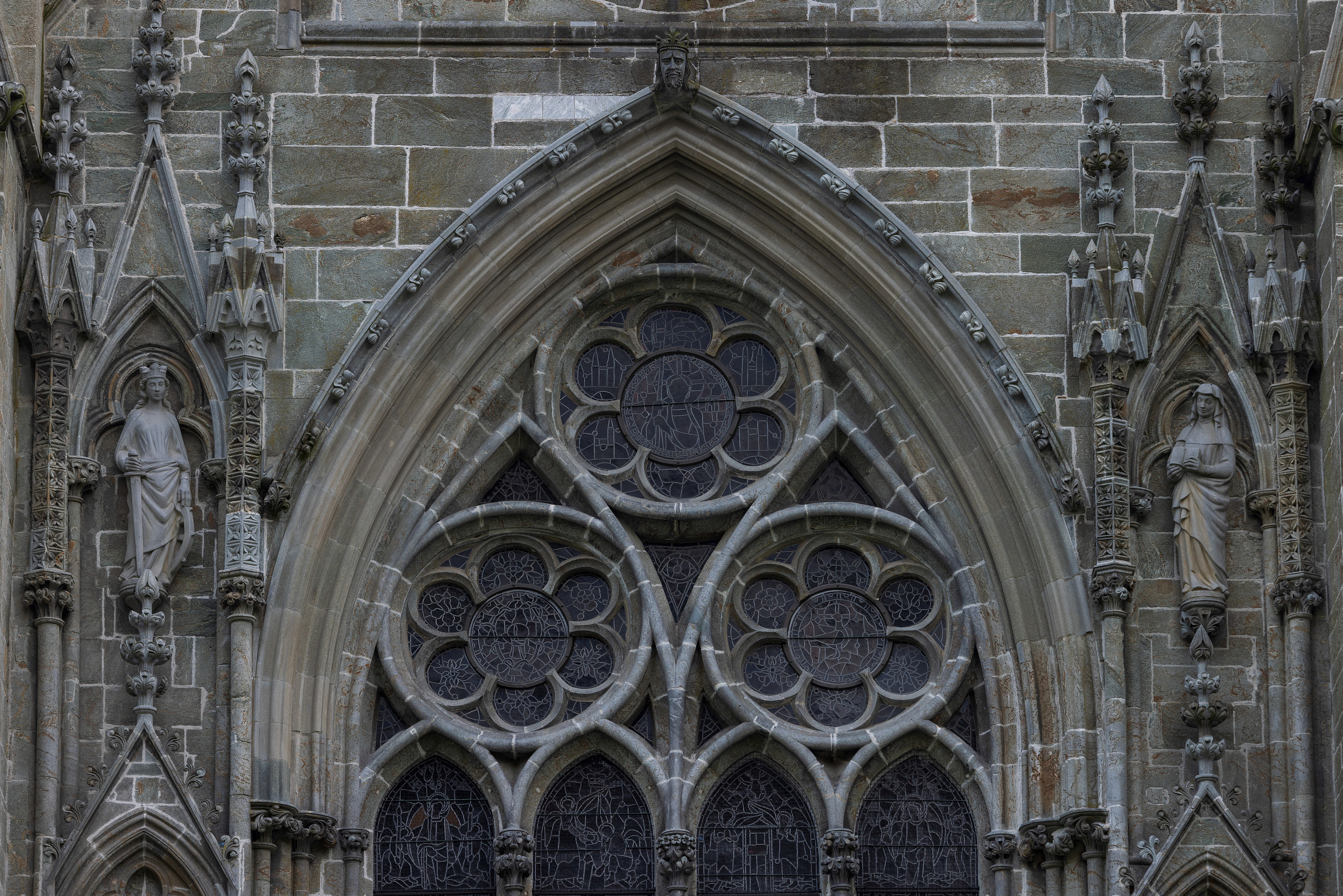 Detaljbilde av en gotisk katedralfasade med spissbuer, utsmykkede sprosser, glassmalerier og steinstatuer plassert i nisjer.