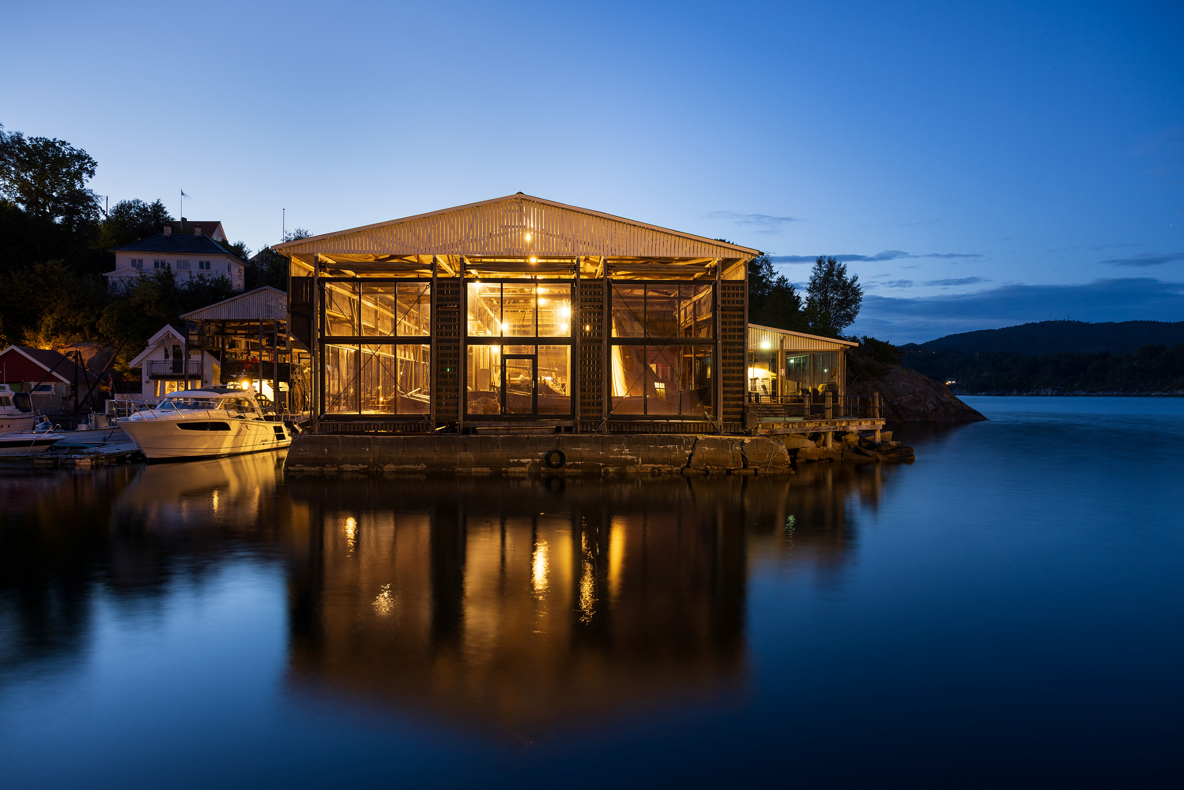 Et notheng med glassvegger som lyser opp i skumringen og speiler seg i den rolige havnefronten med båter som ligger fortøyd like ved med åser i bakgrunnen.
