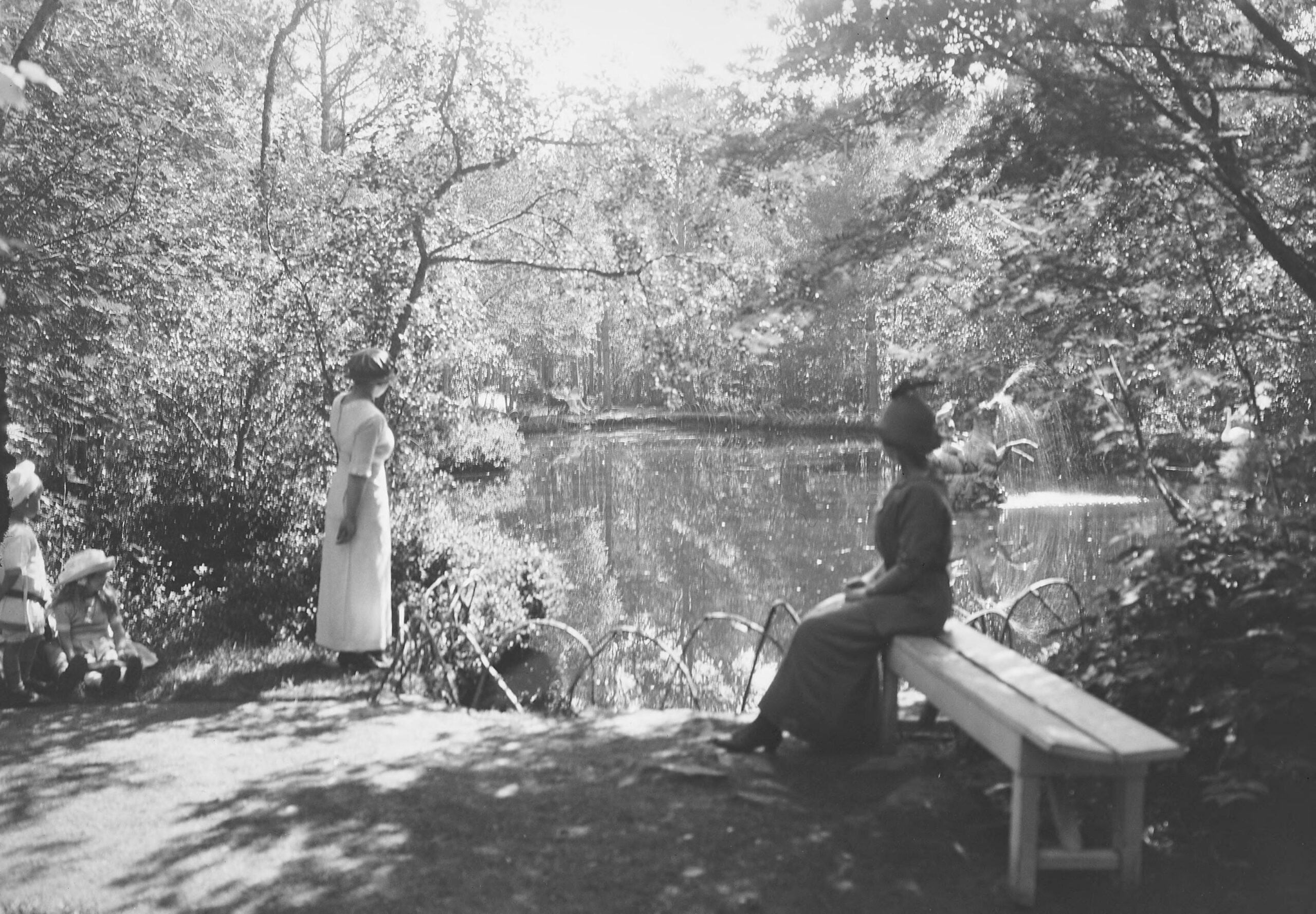 Svart-hvitt-foto av kvinner og barn ved et tjern i et skogsområde; en kvinne sitter på en benk, mens andre står eller sitter ved vannet.