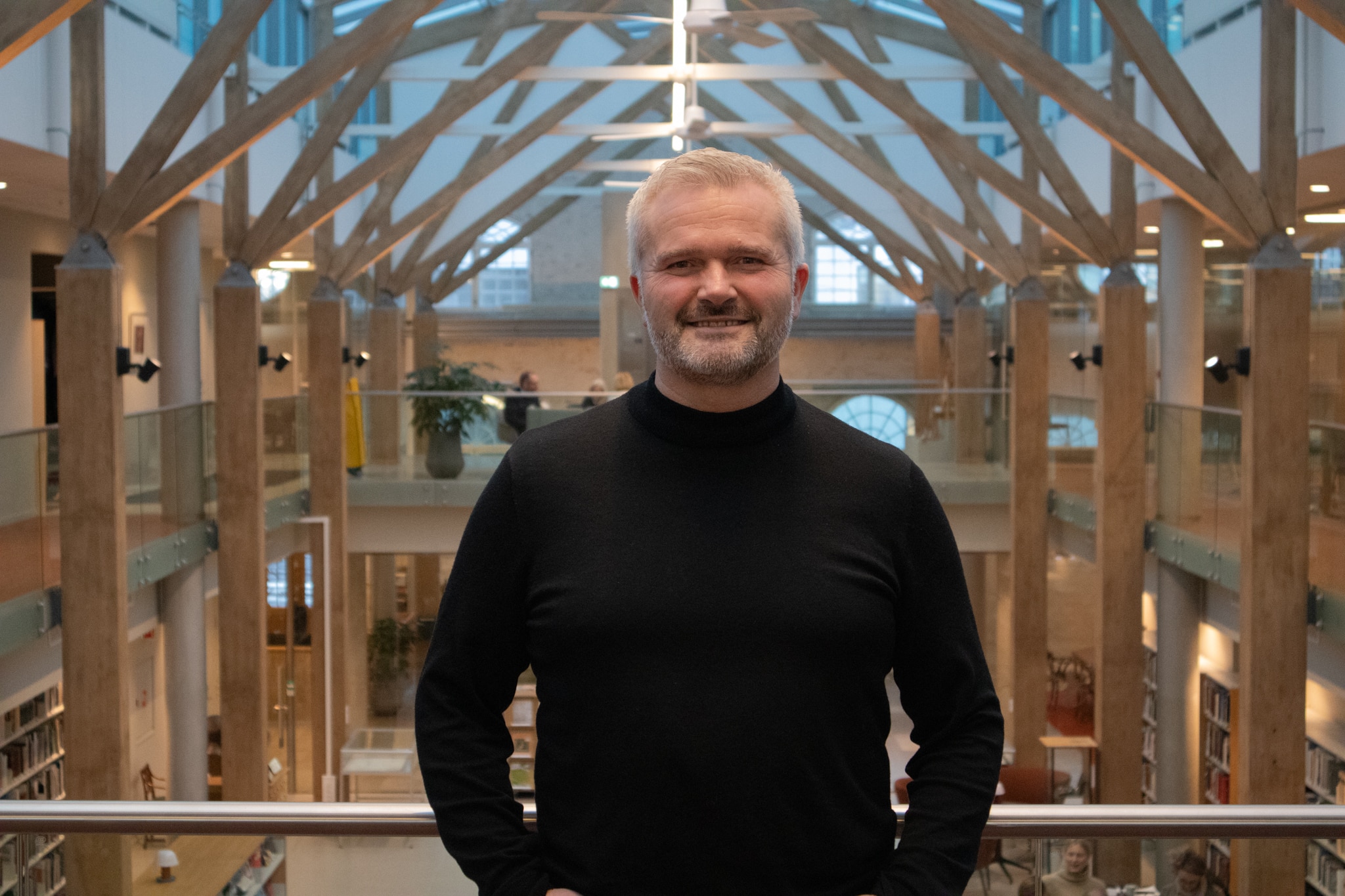 En mann med grått hår og skjegg, iført svart rullekrage, står innendørs i et moderne bibliotek med synlige bjelker og bokhyller i bakgrunnen.