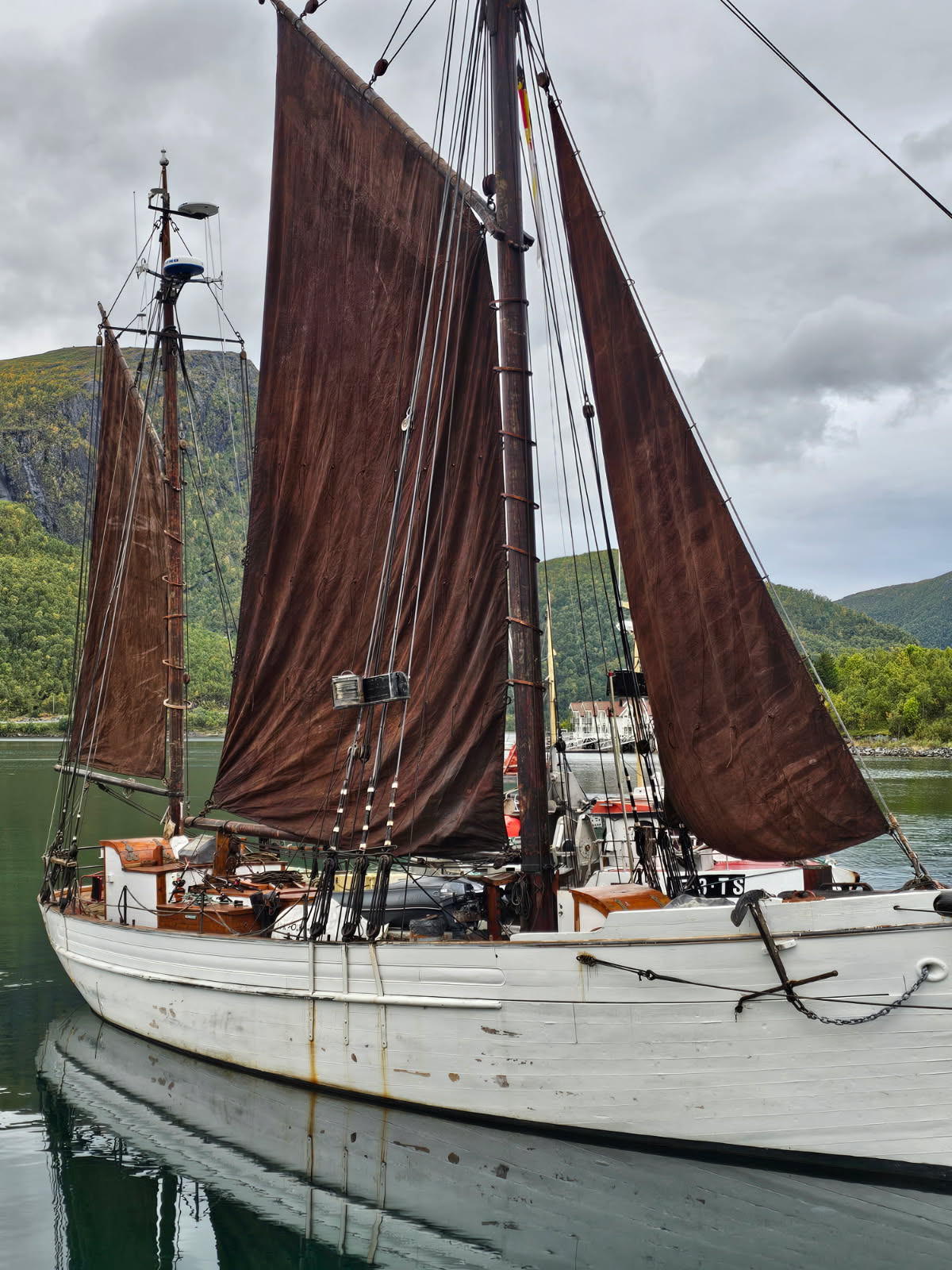 En hvit seilbåt i tre med brune seil ligger til kai på stille vann, omgitt av grønne åser og overskyet himmel.