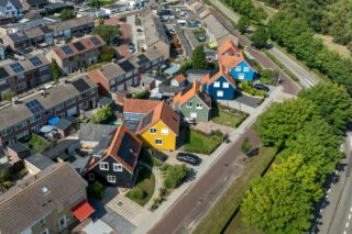 Flyfoto av et boligområde i Nederland med fargerike hus, inkludert freder 19 norske hus, solcellepaneler på takene, trær og parkerte biler langs gaten.